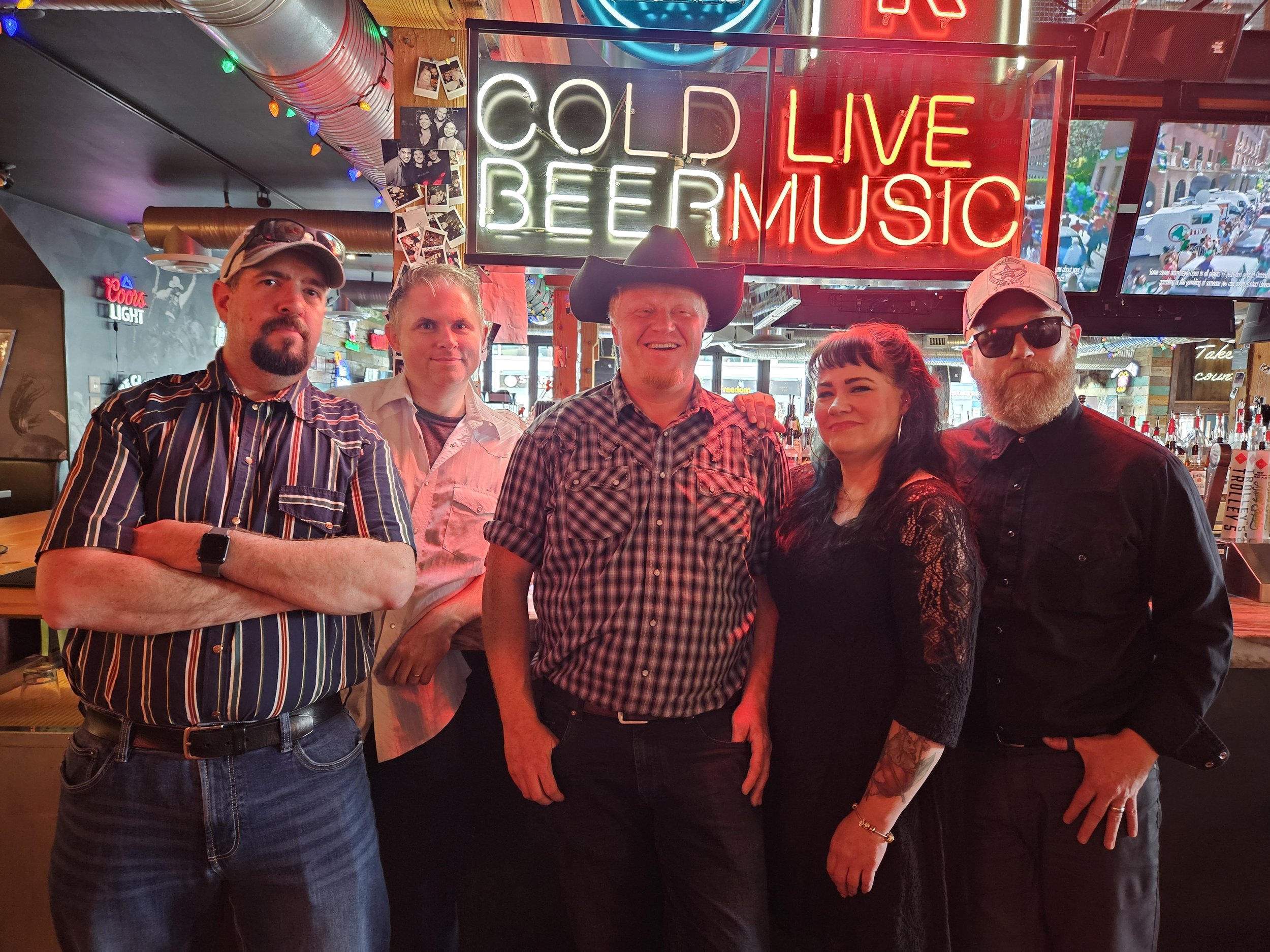 Group of five people standing in a bar with a neon sign saying Cold Beer Live Music